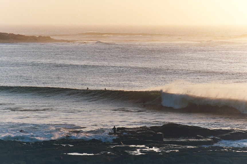 A beach at sunset with surfers in the water and a small rocky ridge on the left hand side of the bay