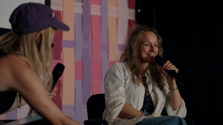 Samantha Morton wearing a white shirt, holding a microphone close to her face on stage - the background is purple, peach and pink ribbons