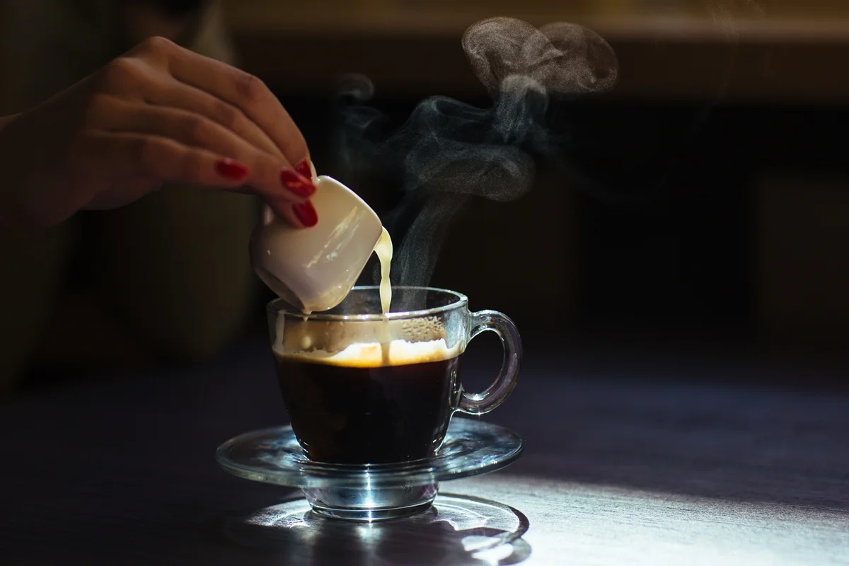 Foto colorida de uma mão feminina derrubando café quente em uma xícará que está enfatizada pela luz do ambiente - Metrópoles.