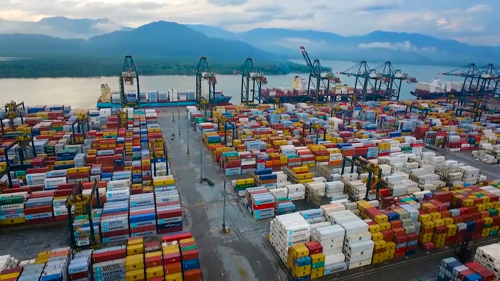 Vista aérea de um porto de contêineres, com numerosos contêineres de diferentes cores (vermelho, azul, amarelo, verde) organizados em fileiras. Ao fundo, gruas e navios estão ancorados, com montanhas ao longe sob um céu nublado.