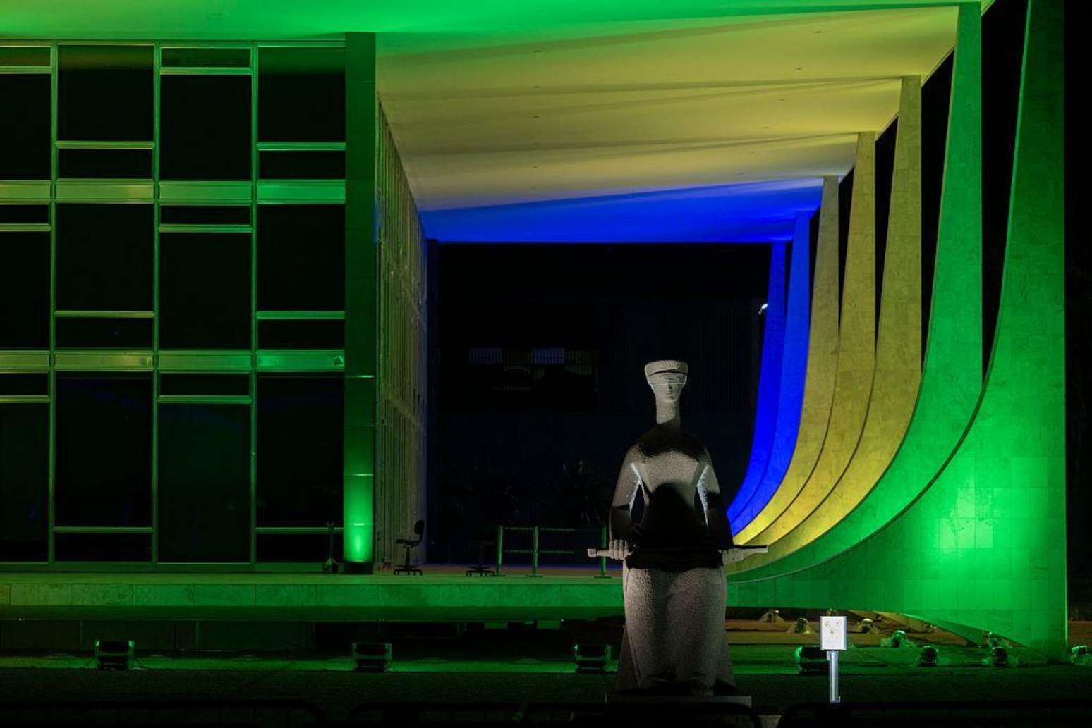 Foto noturna do STF, com estátua da Justiça na frente e colunas em cores da bandeira brasileira
