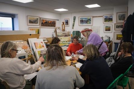Group of people around a table surrounded by paintings and artwork, one in lilac standing up at the far end to help a seated person dressed in red.