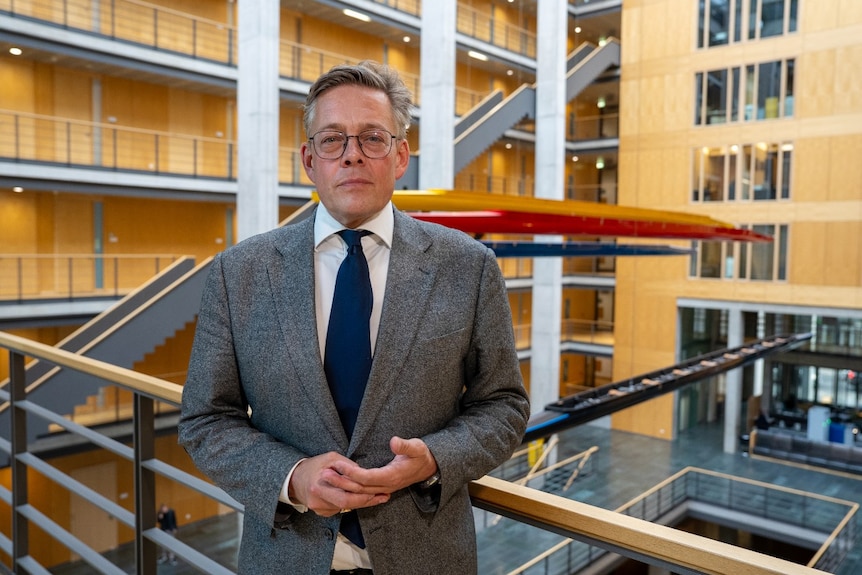 A man in a suit looks at the camera with a neutral expression. He is standing in the atrium of a large building.