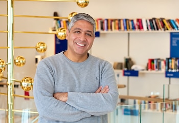 Hombre de cabello canoso y suéter gris posa con los brazos cruzados frente a un fondo con libros y una escultura metálica dorada