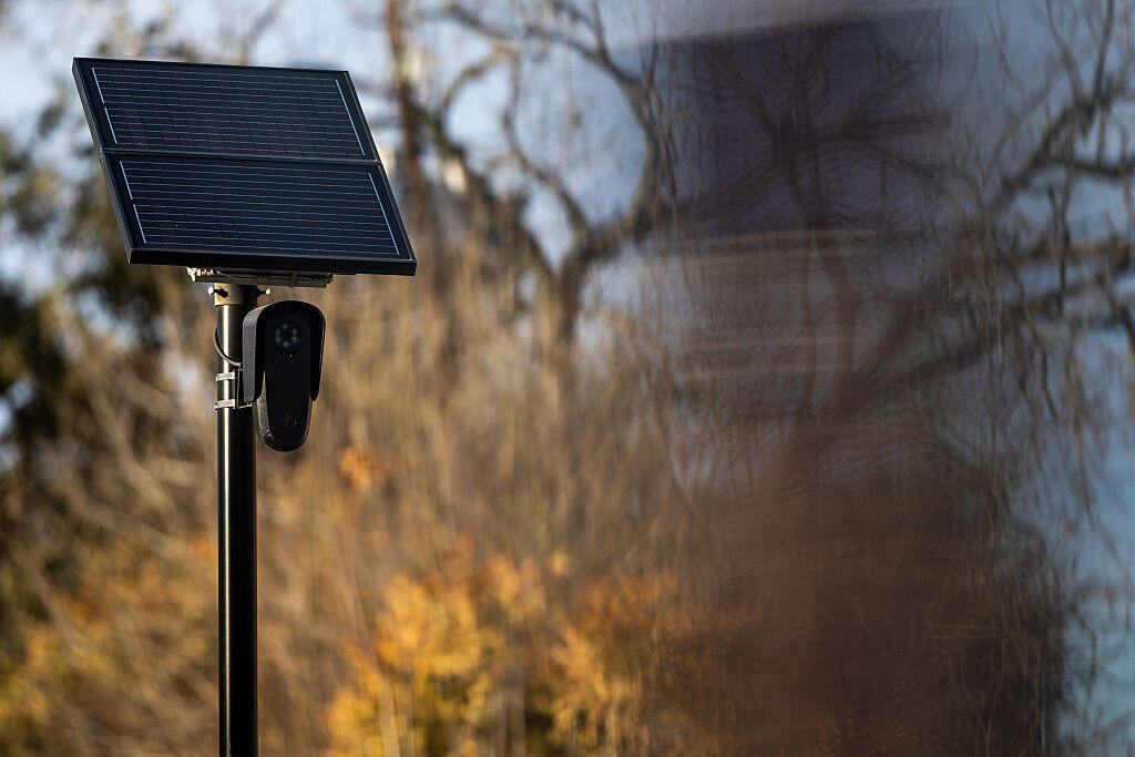 A Flock surveillance camera on a post by trees with a large solar panel connected.